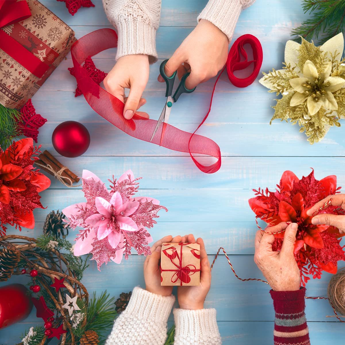Hands crafting Christmas decorations with glitter poinsettias, ribbon, and a gift box on a blue wooden table.