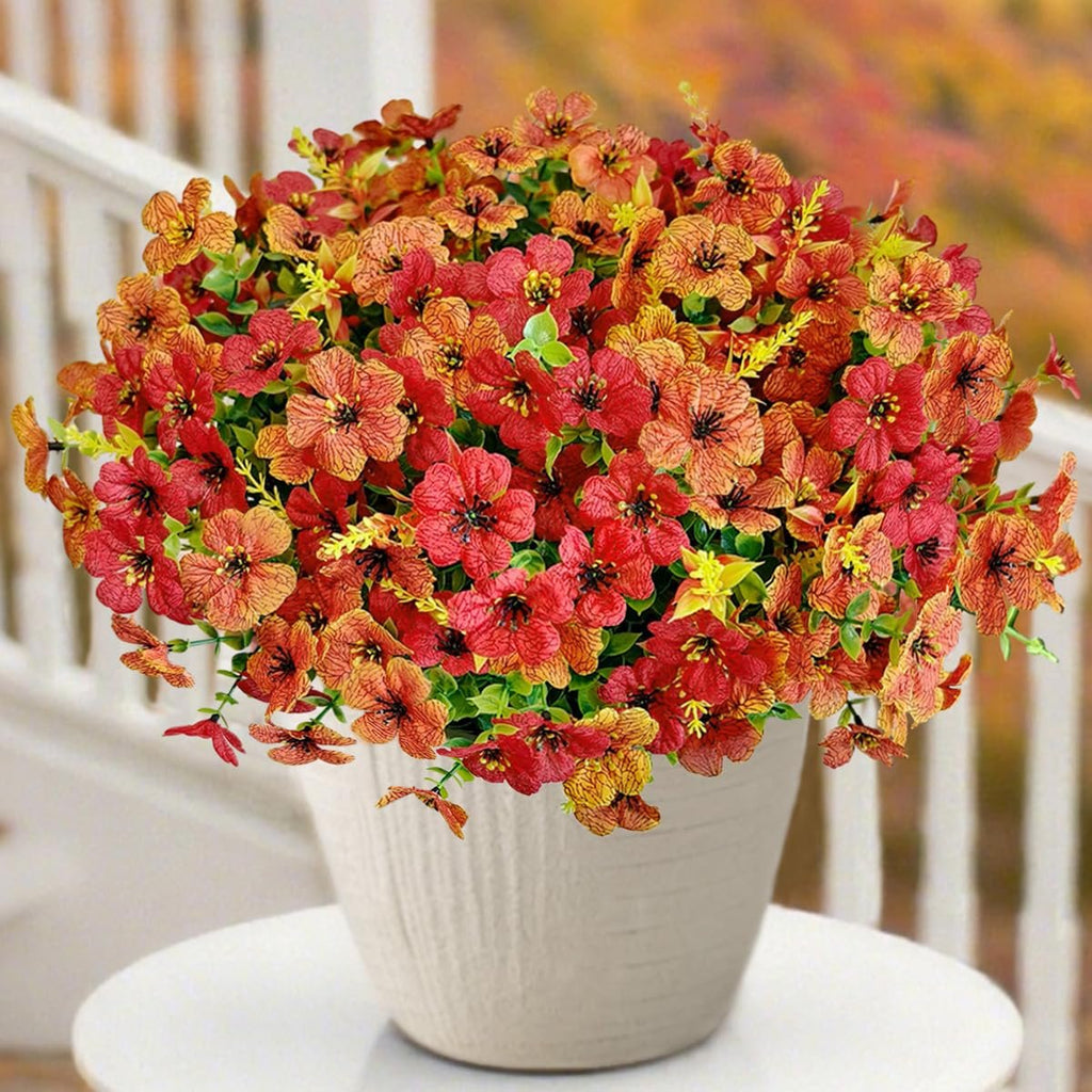 Half sphere of artificial orange and red mums in a white pot, displayed outdoors on a table with a railing and blurred autumn foliage in the background.