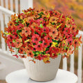 Half sphere of artificial orange and red mums in a white pot, displayed outdoors on a table with a railing and blurred autumn foliage in the background.