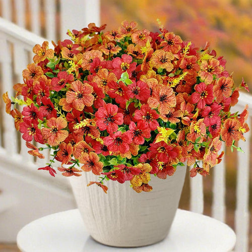 Half sphere of artificial orange and red mums in a white pot, displayed outdoors on a table with a railing and blurred autumn foliage in the background.