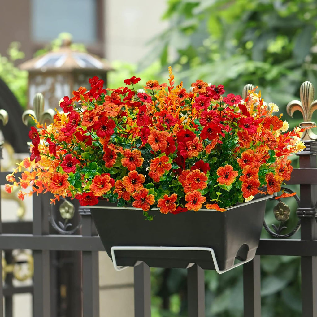 Artificial mums in a planter box hanging on a black metal fence, featuring red and orange flowers with green foliage.