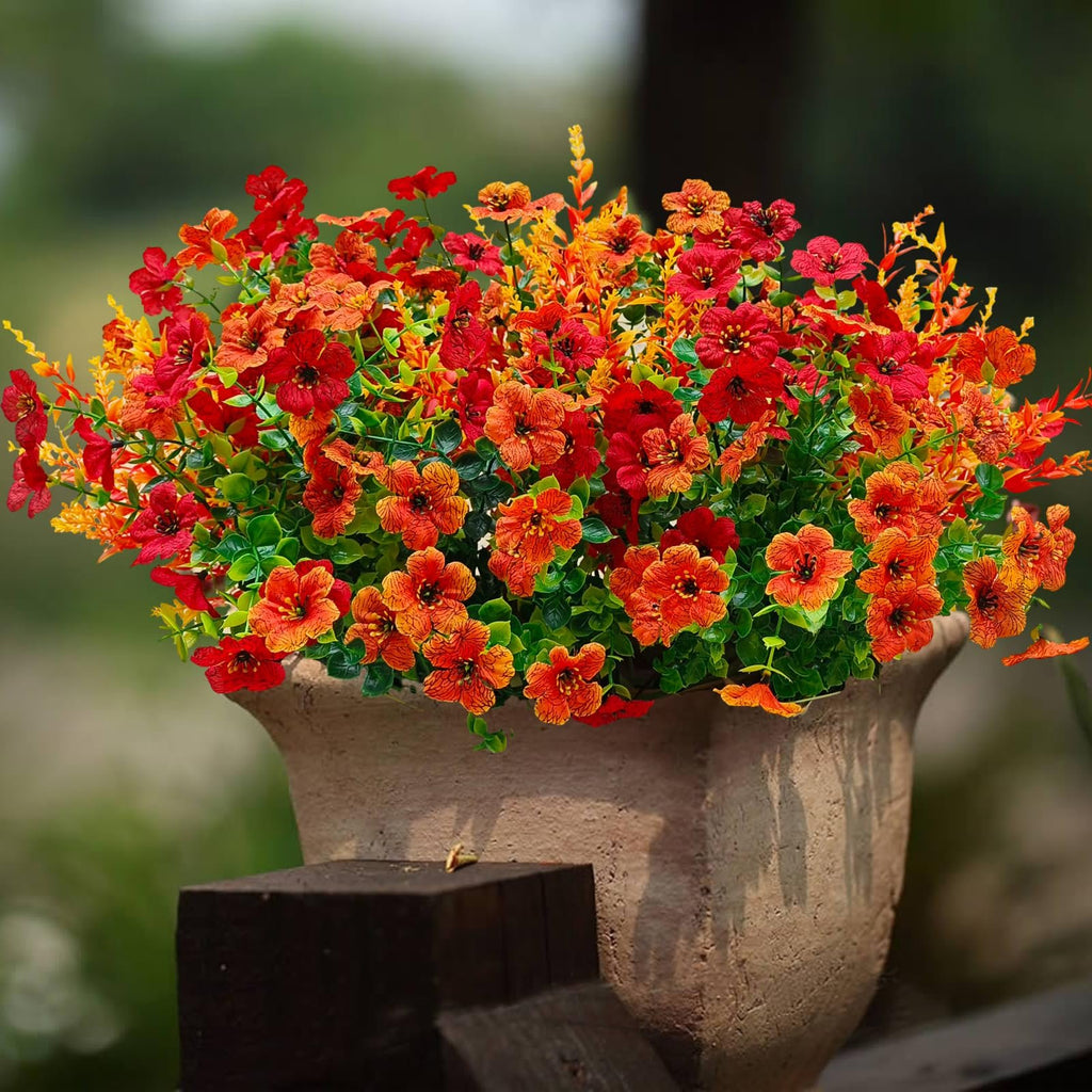 Artificial orange and red mums in a terracotta pot on a wooden post, with a blurred green background.