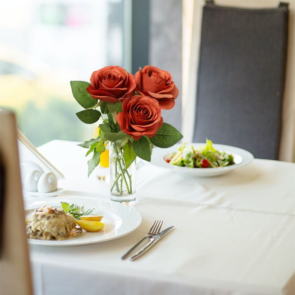 Three burnt orange artificial silk roses in a glass vase on a table set for a meal.