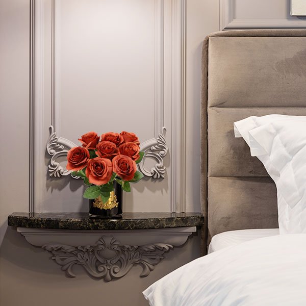 Bouquet of burnt orange artificial silk roses in a black vase with gold trim on a shelf next to a bed.