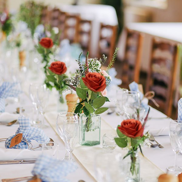 Burnt orange artificial silk roses with green leaves arranged in small glass vases on a wedding reception table setting.