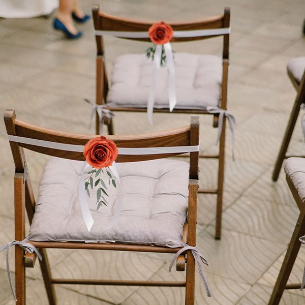 Two wooden chairs decorated with burnt orange silk roses and white ribbons for a wedding ceremony