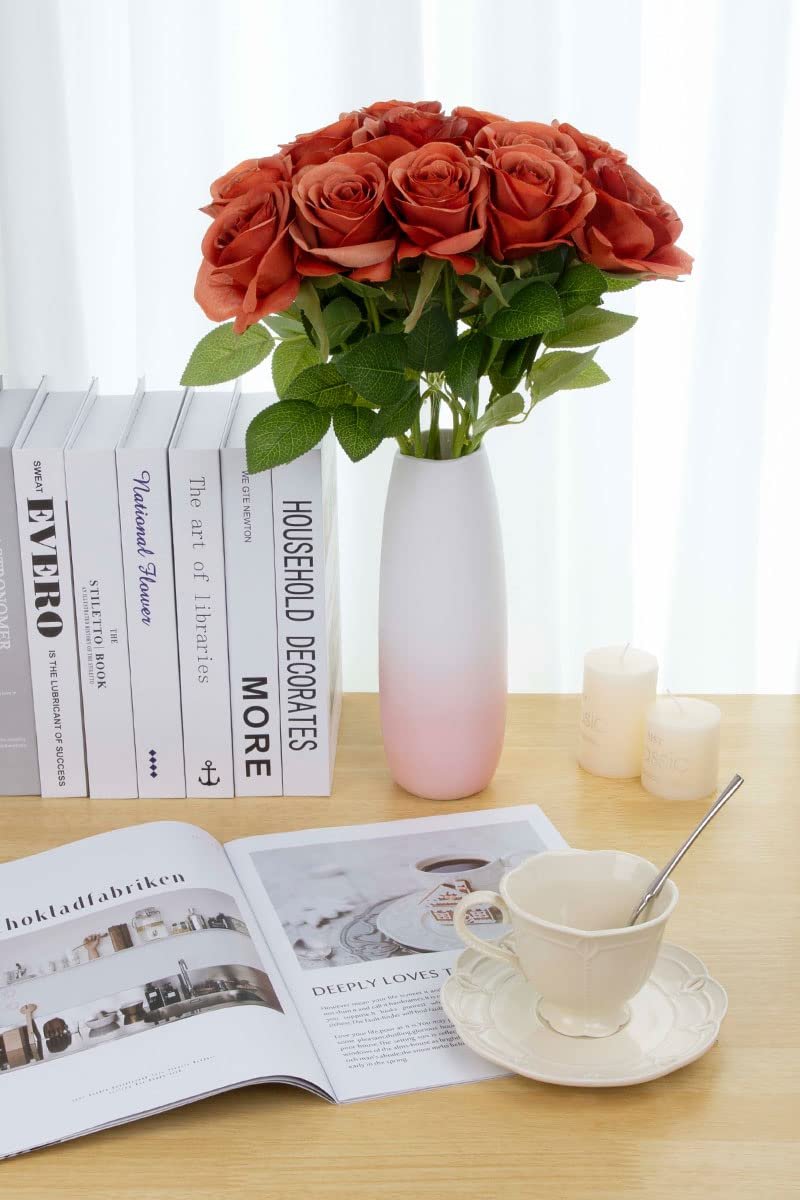 A dozen burnt orange artificial silk roses in a white and pink gradient vase, arranged on a wooden table with books and a teacup.