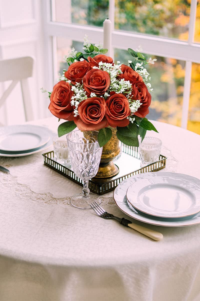 Burnt orange artificial silk roses in a gold vase on a table setting with plates and a wine glass.