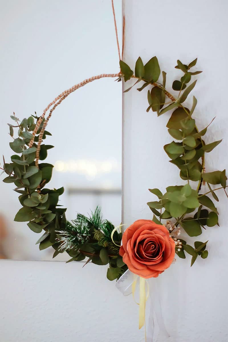 Burnt orange artificial rose wreath with eucalyptus leaves and pine sprigs hanging on a white wall, reflecting lights.