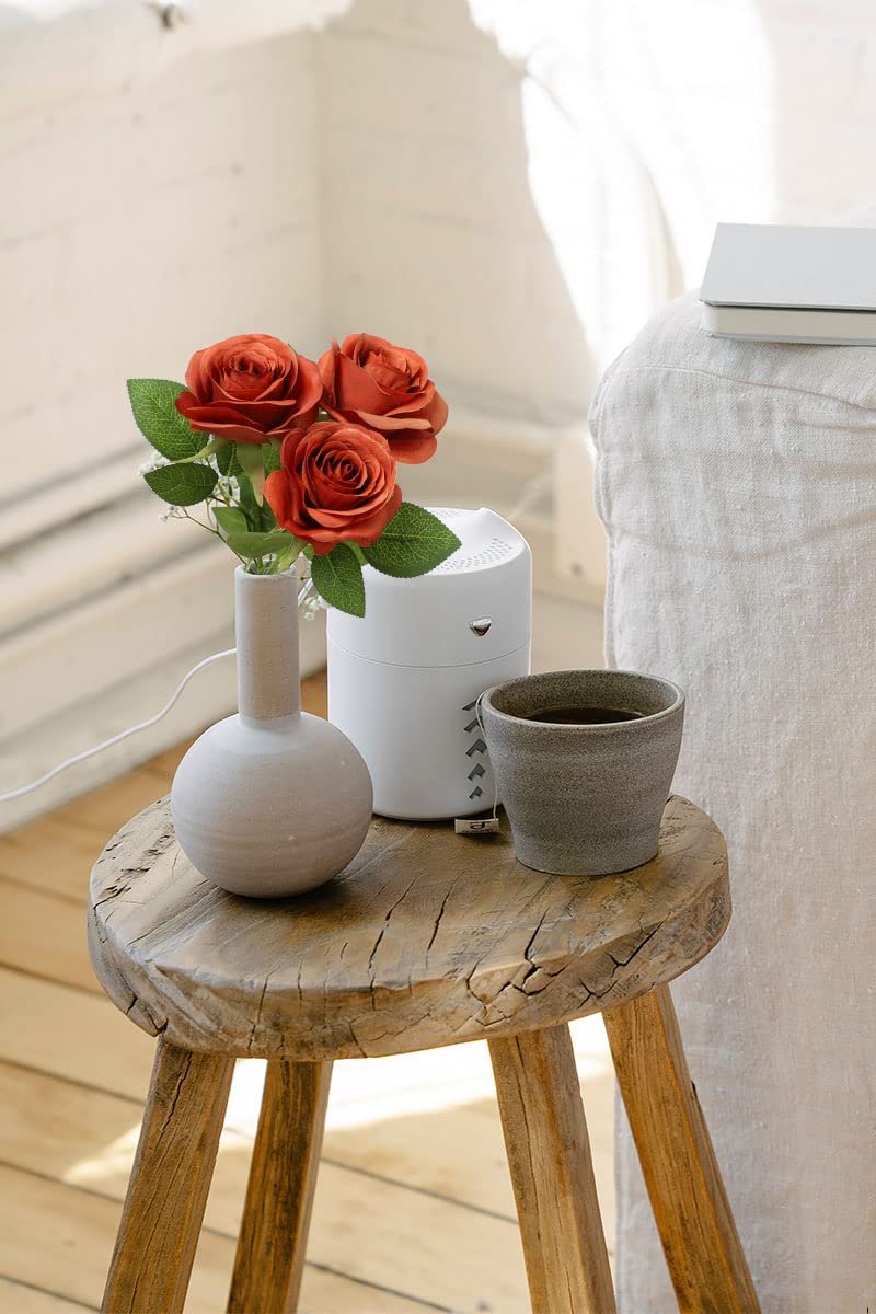 Three burnt orange artificial roses in a vase on a rustic wooden stool next to a cup of tea and a white humidifier.