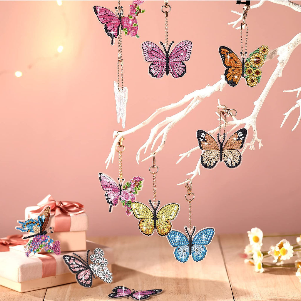 Assortment of diamond painting butterfly keychains hanging from a white branch, with gift boxes and flowers below.