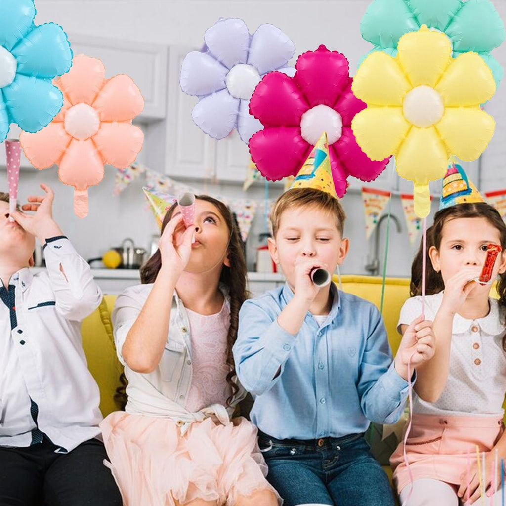 Four children wearing party hats blow party horns with colorful daisy balloons floating above them
