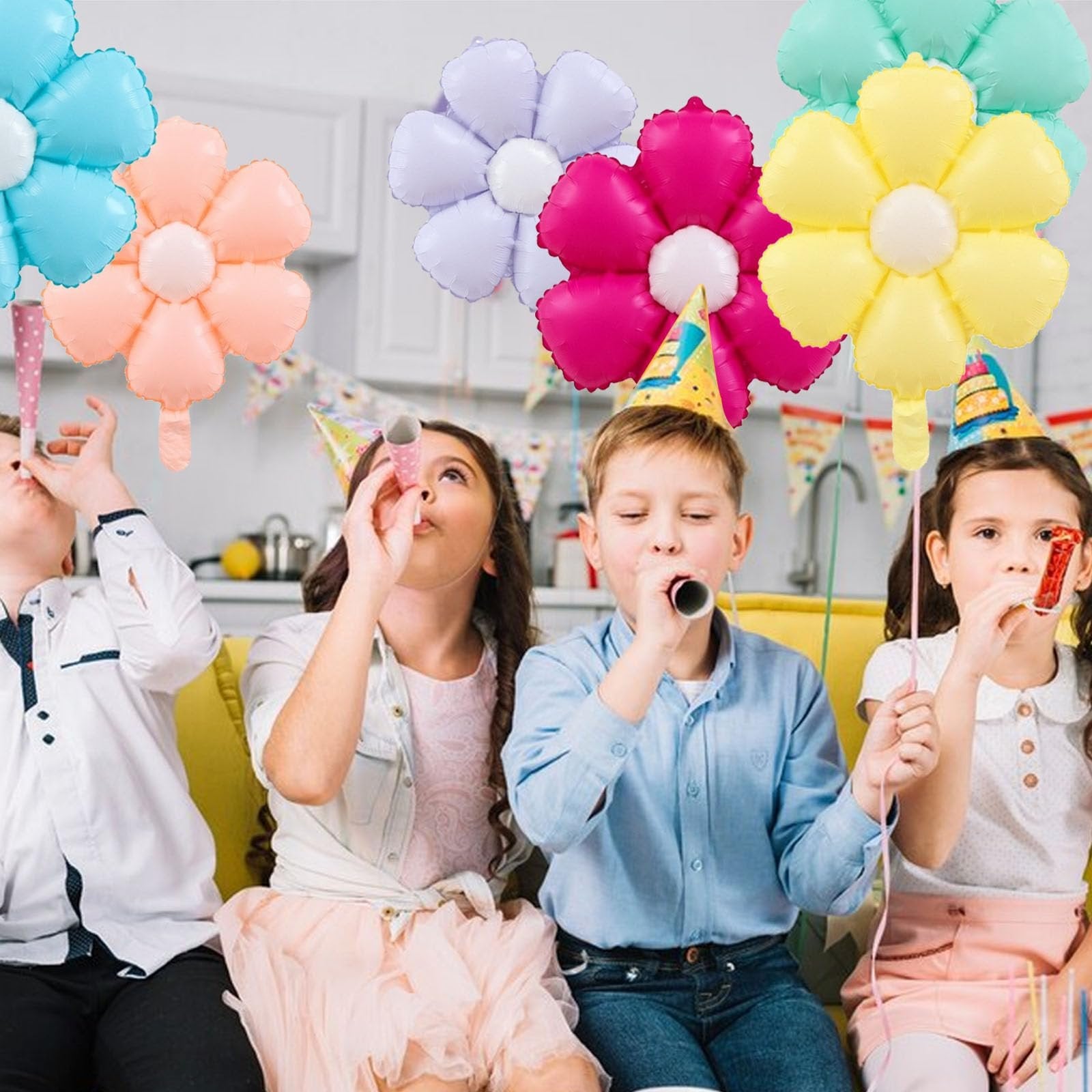 Four children wearing party hats blow party horns with colorful daisy balloons floating above them
