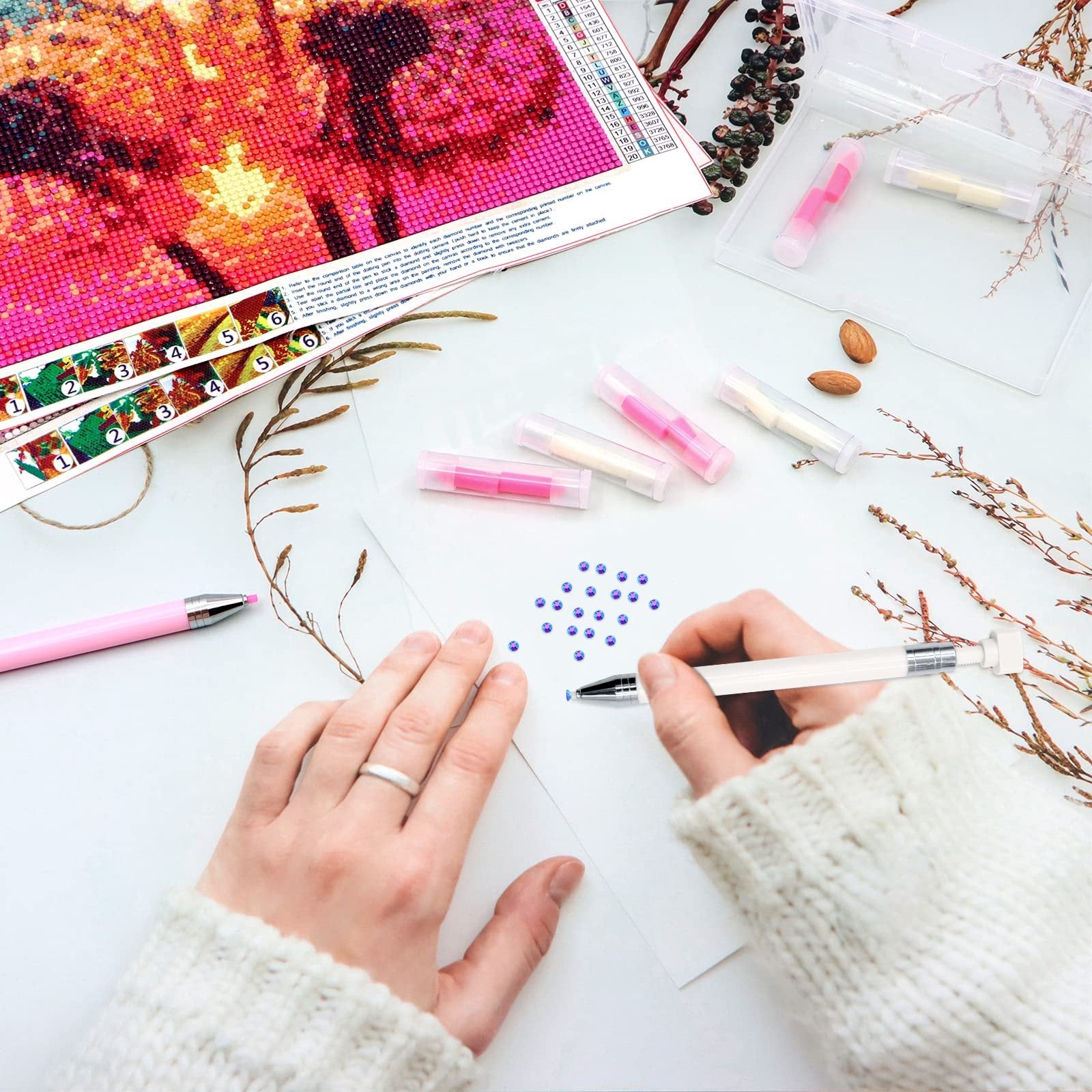 Hands using diamond painting tools to place blue rhinestones on paper, with wax refills and storage box nearby.