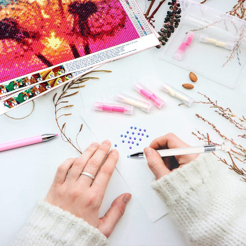 Hands using diamond painting tools to place blue rhinestones on paper, with wax refills and storage box nearby.