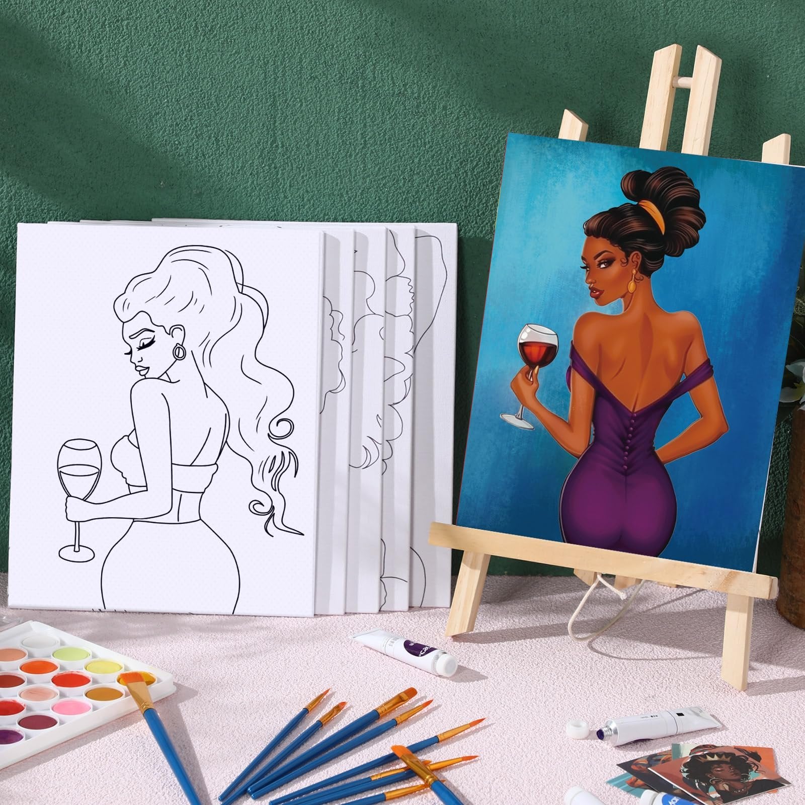 Two pre-drawn canvases for painting, one outlined and one fully painted, with art supplies like brushes and paint tubes on a pink surface.