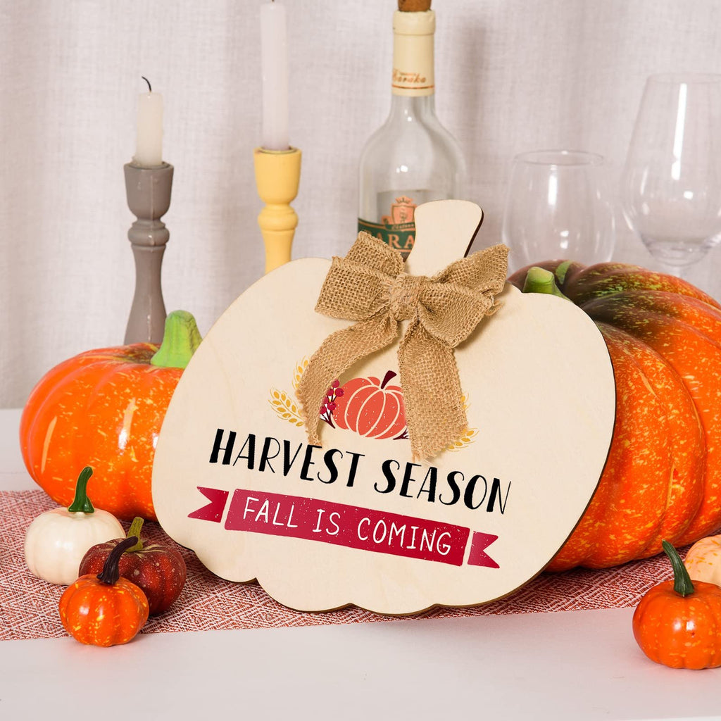 Wooden pumpkin cutout with burlap bow, decorated for fall with text 'HARVEST SEASON FALL IS COMING', surrounded by decorative pumpkins and candles.