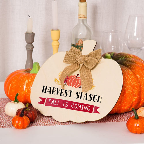 Wooden pumpkin cutout with burlap bow, decorated for fall with text 'HARVEST SEASON FALL IS COMING', surrounded by decorative pumpkins and candles.