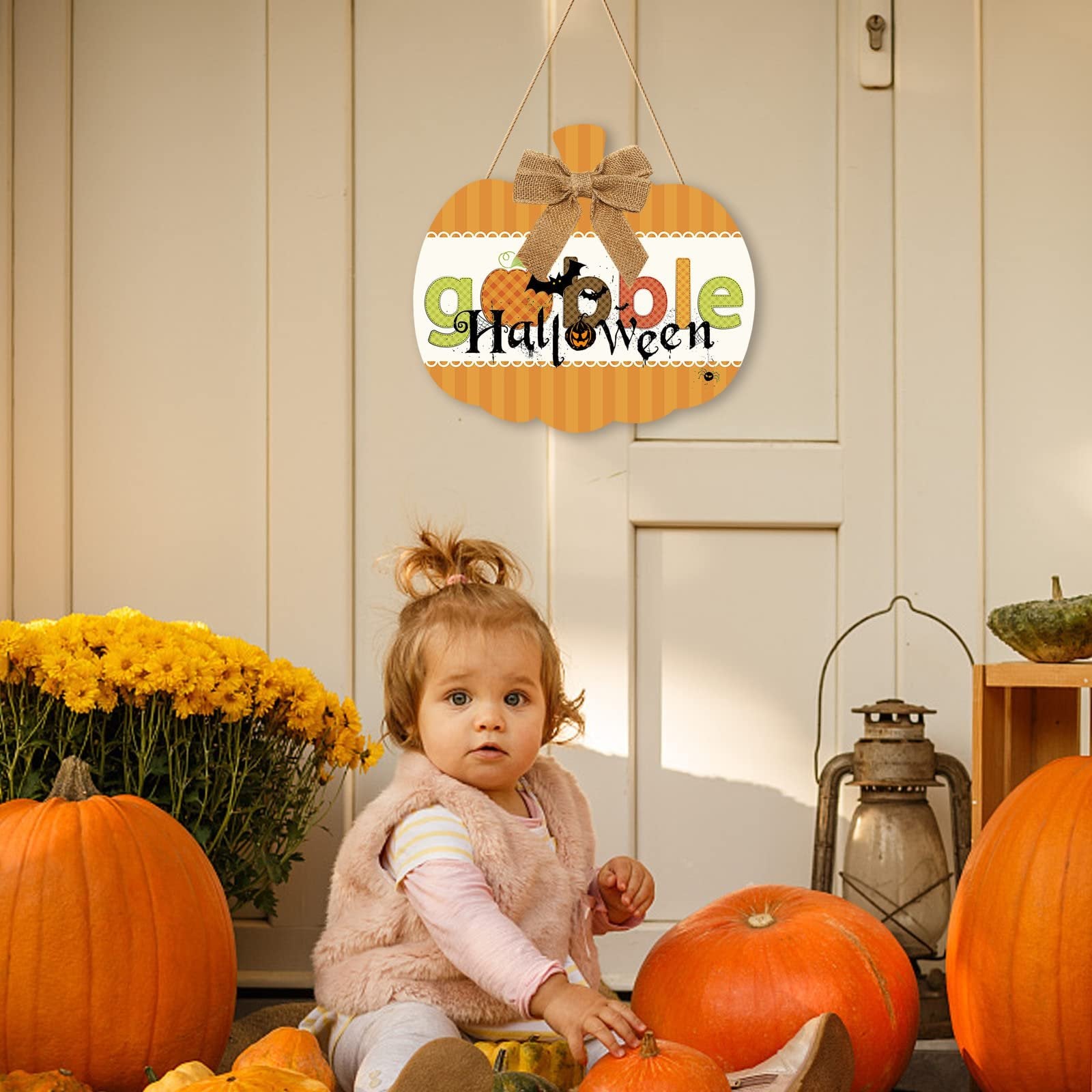 Toddler sitting among pumpkins and fall decor with a hanging wooden pumpkin sign that says 'Gobble Halloween'