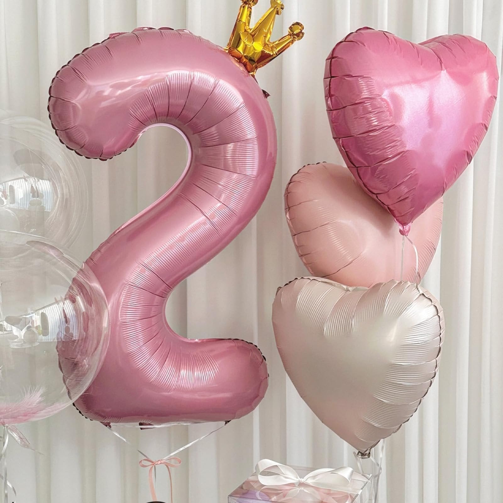 Pink number 2 balloon with gold crown and pink heart balloons against a white curtain background.