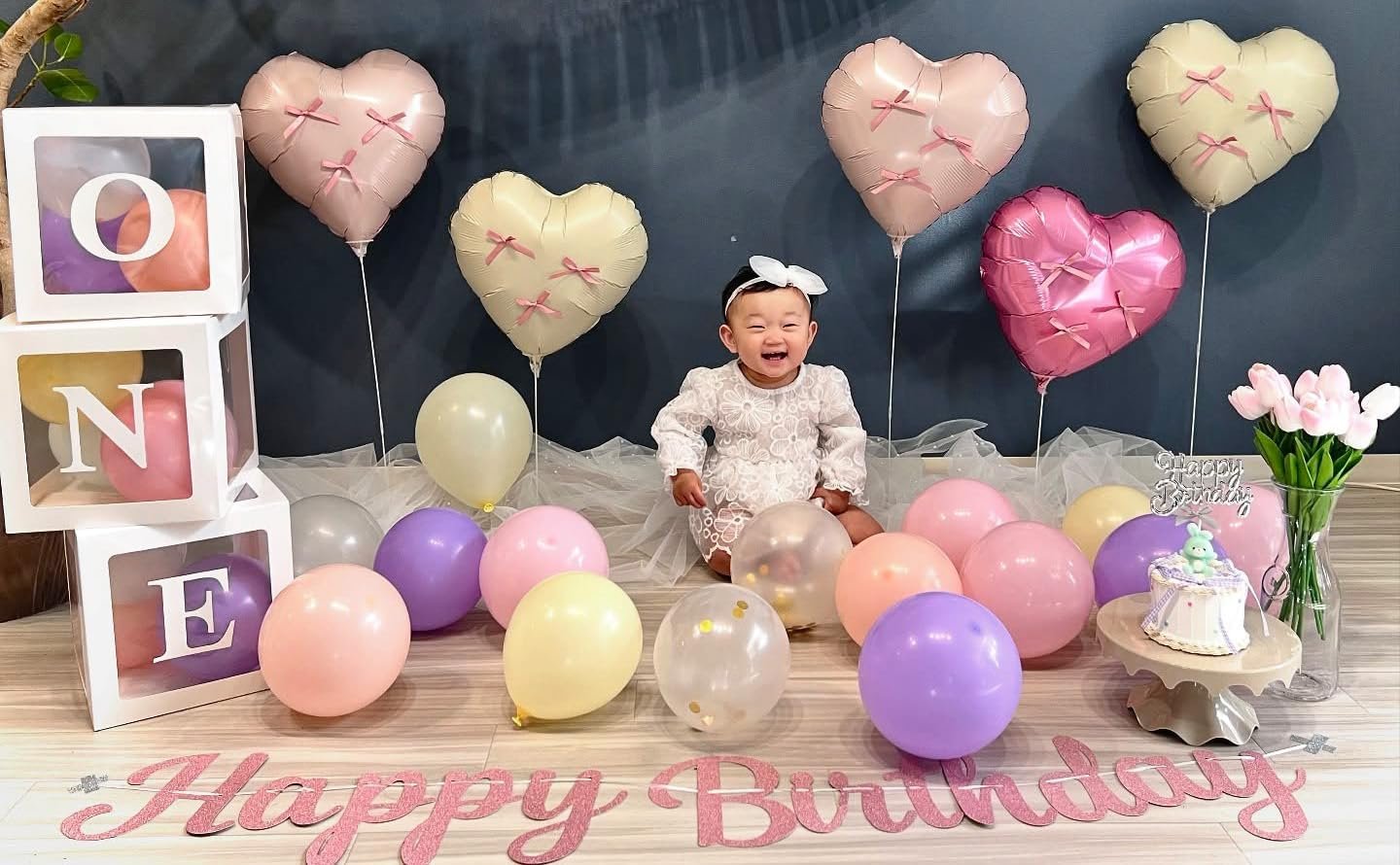Baby girl sitting among heart-shaped balloons and a birthday cake, with 'Happy Birthday' banner.