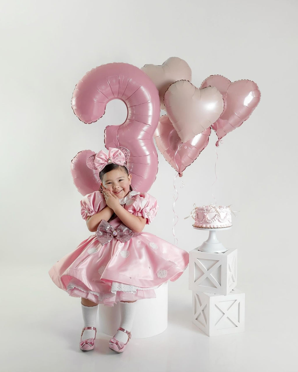 Girl in pink dress with bow headband and heart balloons, celebrating 3rd birthday with cake