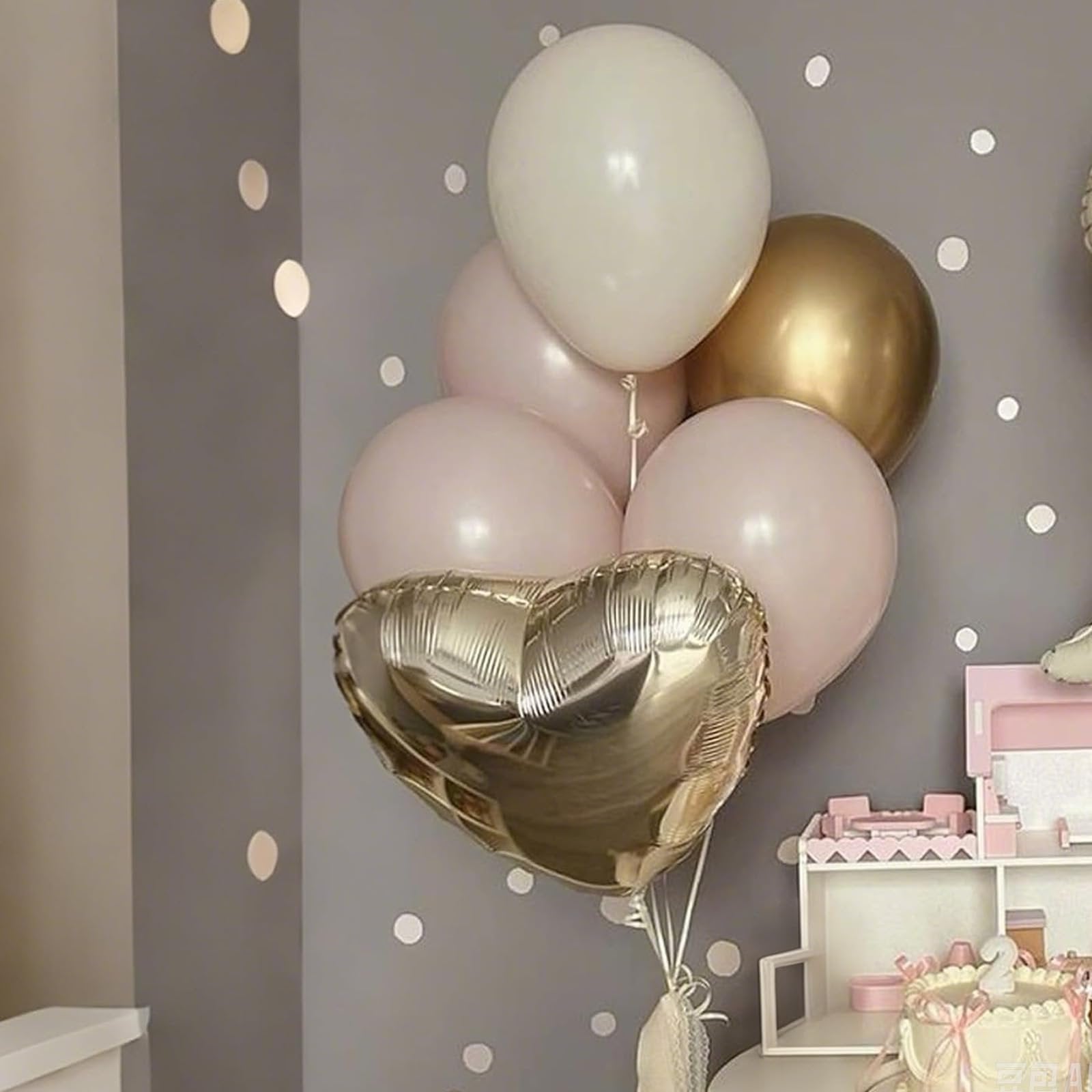 Champagne gold and cream heart and round balloons arranged in a bouquet, with a pink cake and toy kitchen in the background.