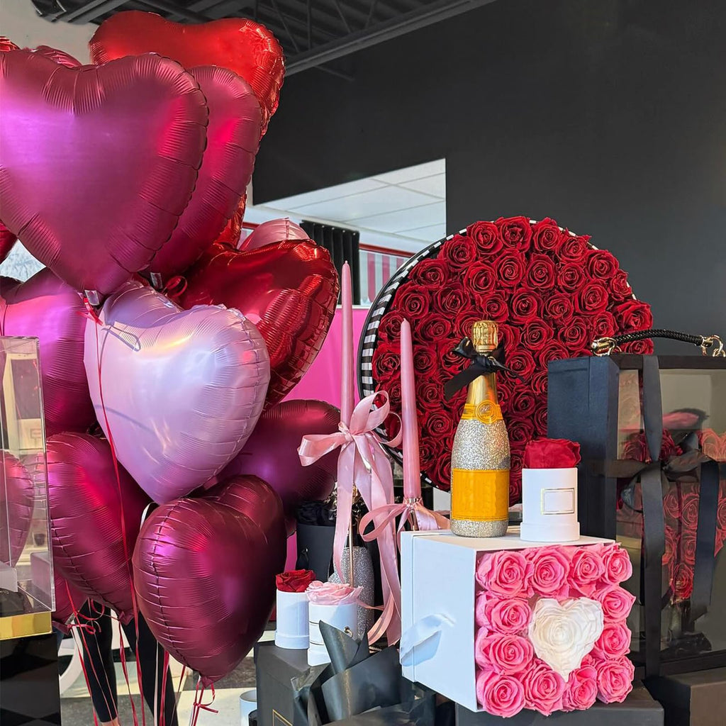 Assortment of heart-shaped foil balloons in red, pink, and rose gold, with a bouquet of red roses and a glittery champagne bottle.