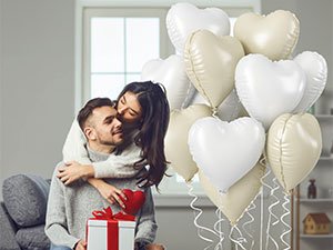Couple embracing with a gift box and a bouquet of pink and white heart-shaped foil balloons