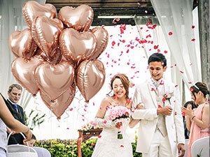 A bride and groom walking down an aisle with rose petals falling, holding a bouquet of pink roses and a bunch of rose gold heart-shaped balloons.