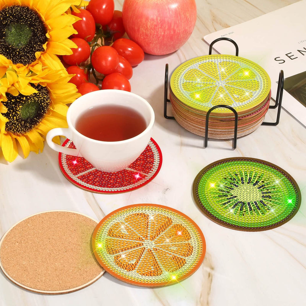 Diamond art coaster kit with fruit designs and holder, displayed with a cup of tea, sunflowers, tomatoes, and an apple.