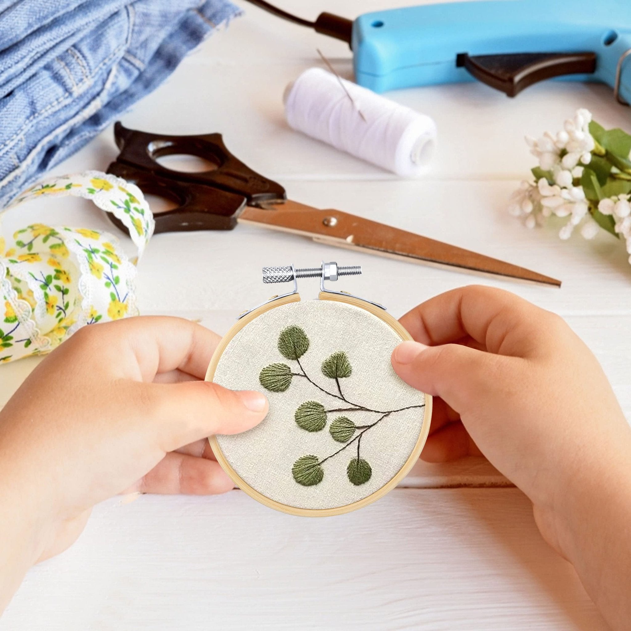 Hands holding a 3-inch bamboo embroidery hoop with green leaf embroidery on light fabric, surrounded by craft supplies.