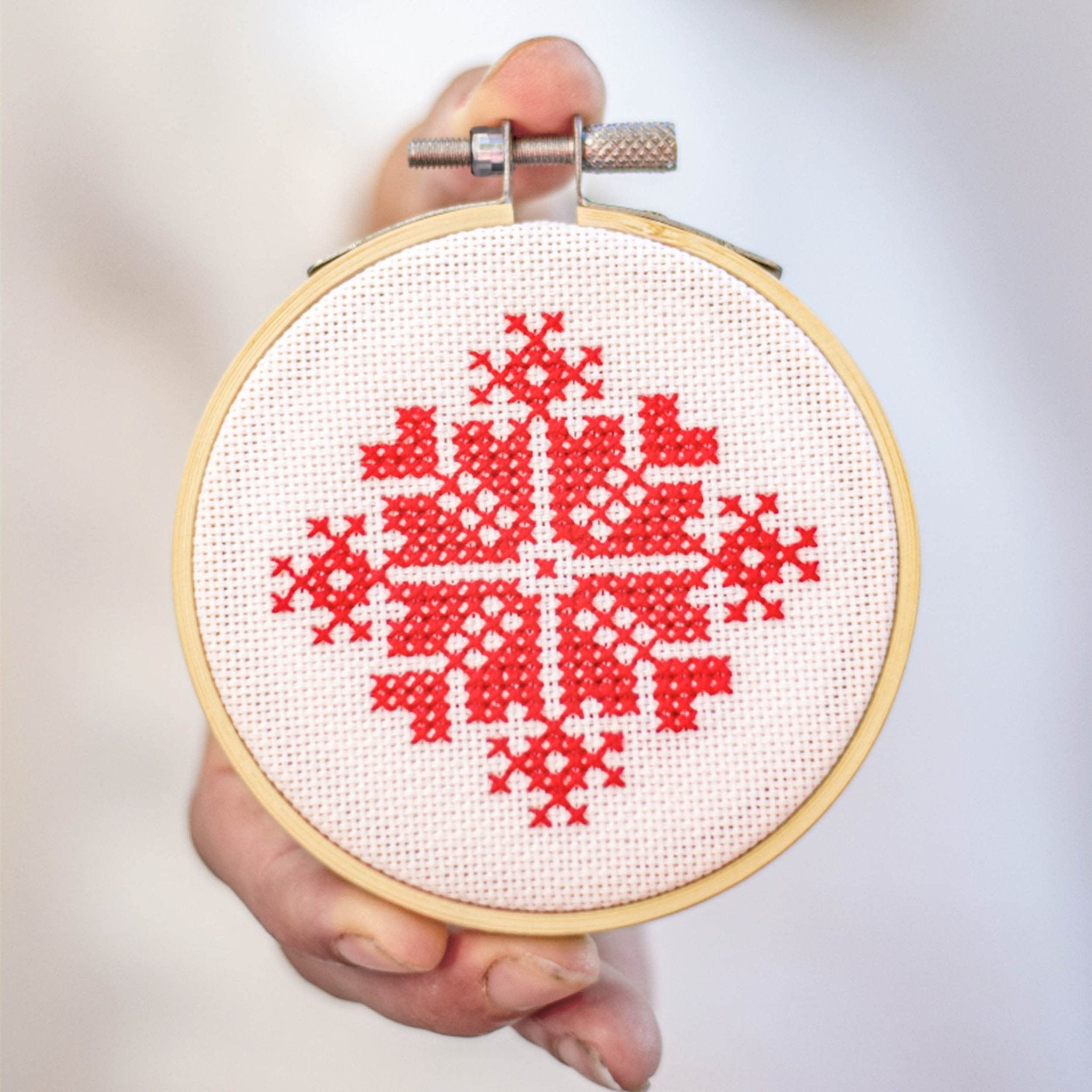 Hand holding a 3-inch bamboo embroidery hoop with white fabric and red cross-stitch pattern