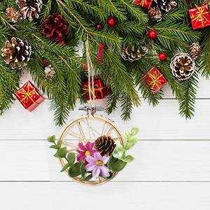 Christmas-themed embroidery hoop decoration with pinecones and flowers hanging from a pine branch against a white wooden background.