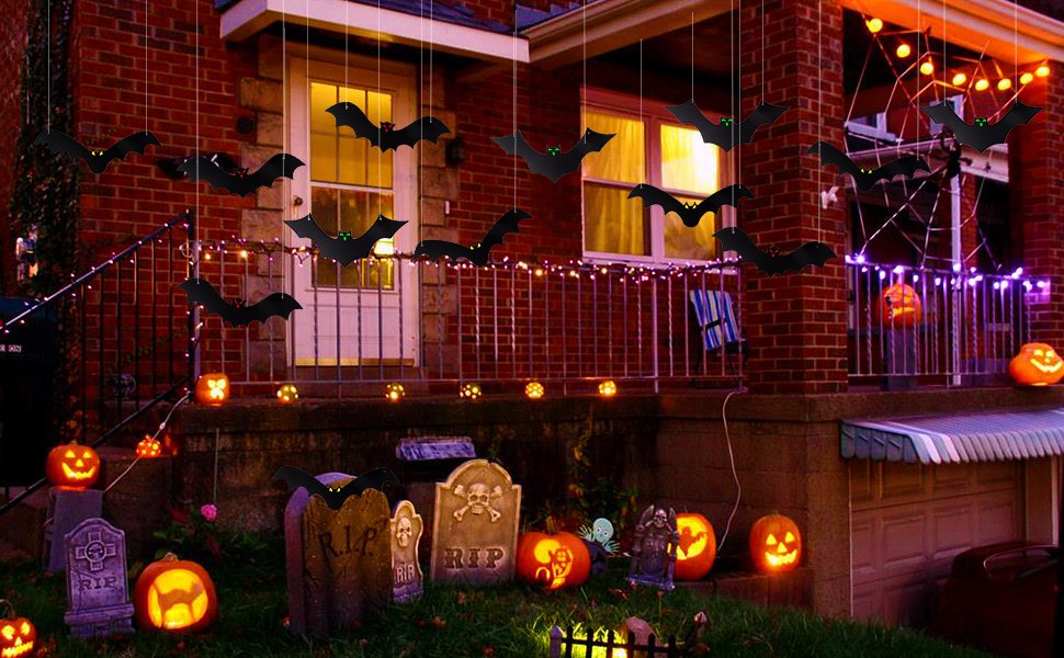 Hanging black bat decorations with glowing eyes on a house porch for Halloween, with pumpkins and tombstones.