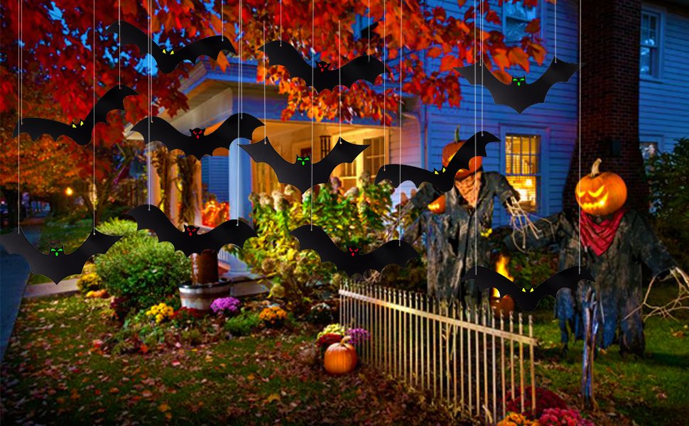 Hanging black bat decorations with glowing eyes displayed outside a house for Halloween, with scarecrows and pumpkins.