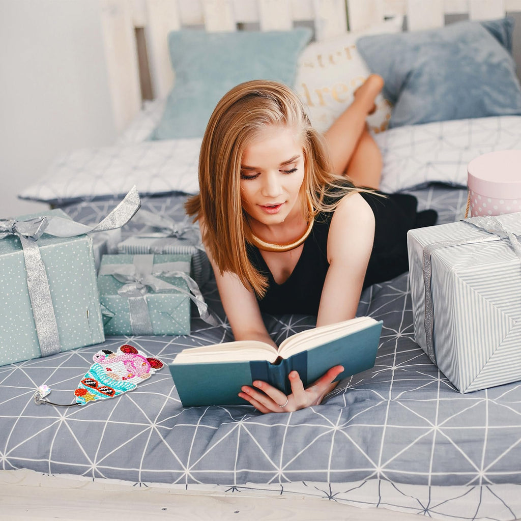 Woman reading a book on a bed surrounded by gift-wrapped boxes, with a diamond-painted gnome bookmark nearby.