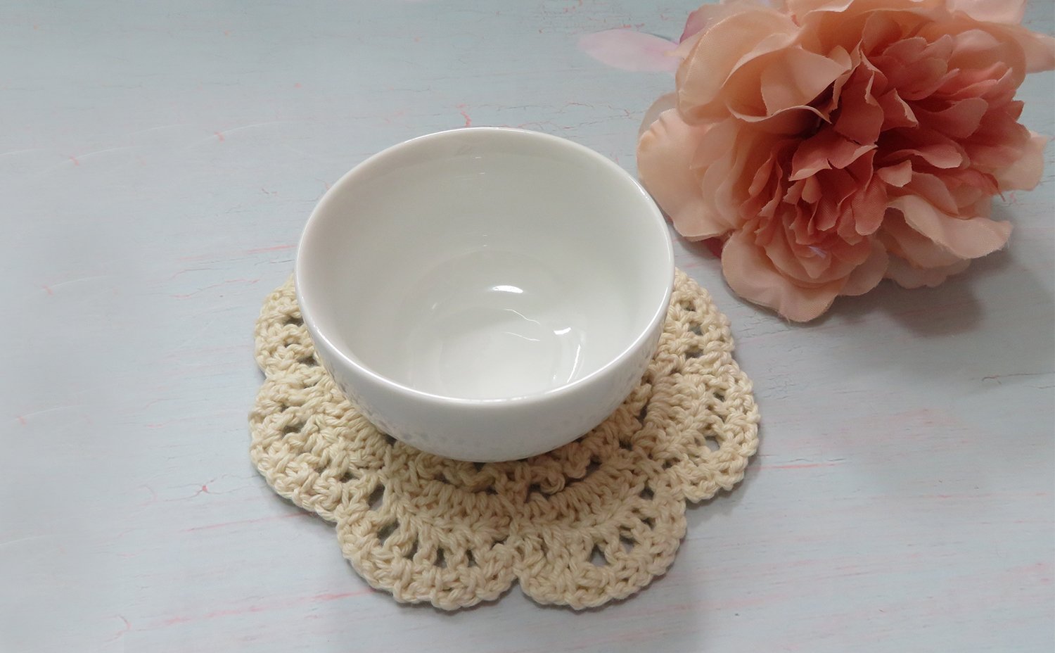 Beige crochet doily with a white bowl on top, next to a pink flower.
