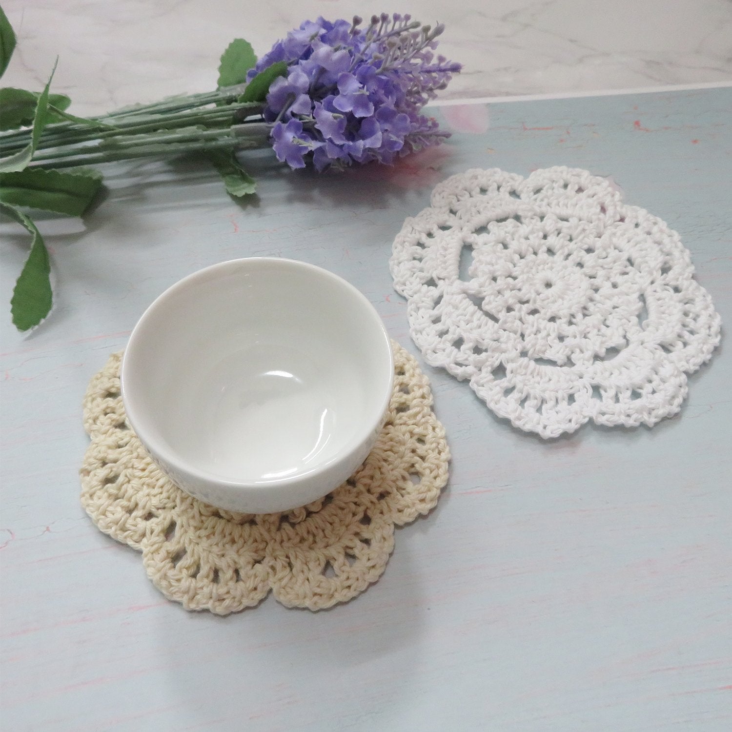 Two handmade crochet doilies, one beige with a white bowl on top, and one white, with lavender flowers in the background.
