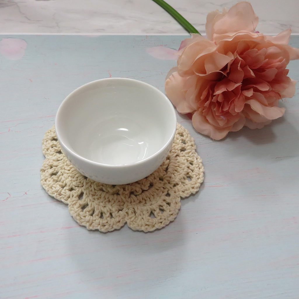 Beige crochet doily with a white bowl on top, next to a pink flower and green stem.