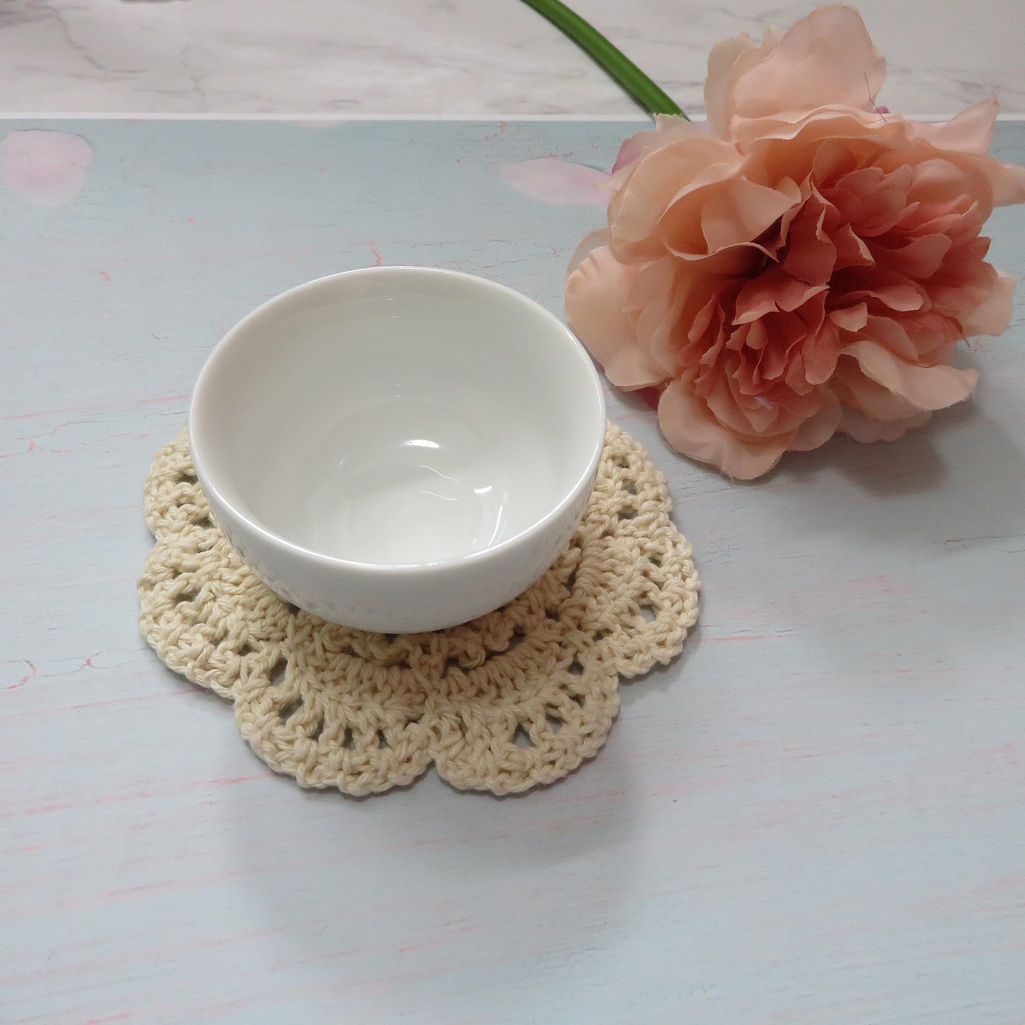 Beige crochet doily with a white bowl on top, next to a pink flower and green stem.