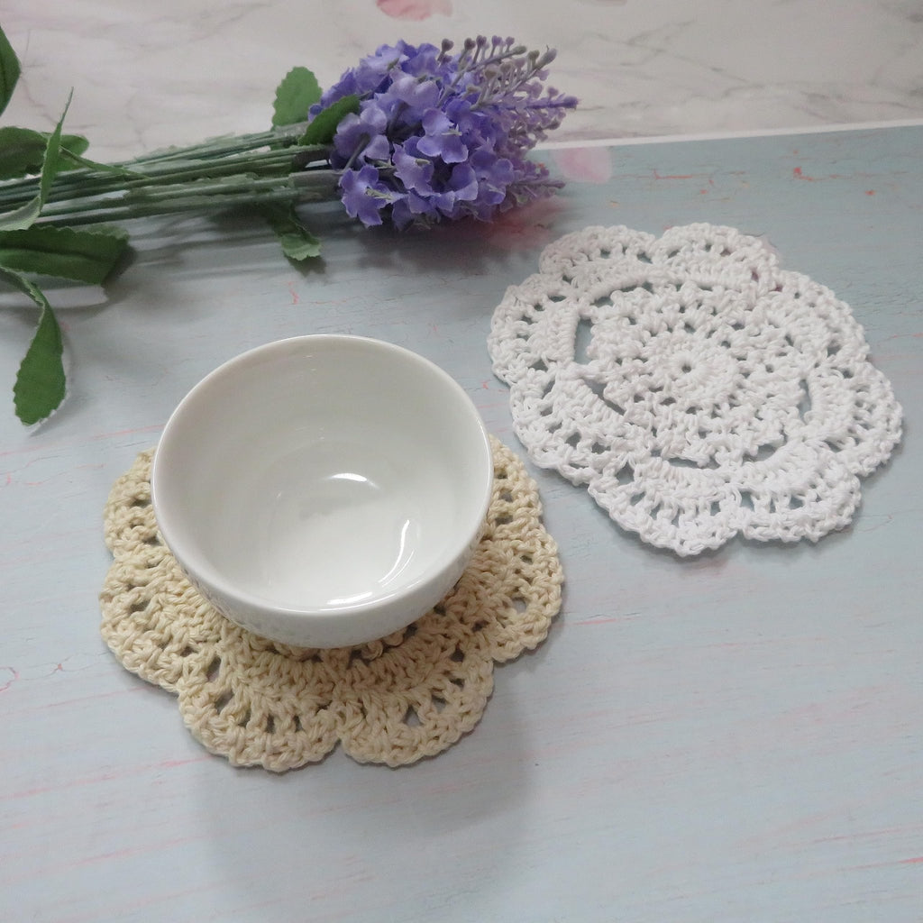 Two handmade crochet doilies, one beige with a white bowl, and one white, with purple flowers and green leaves in the background.