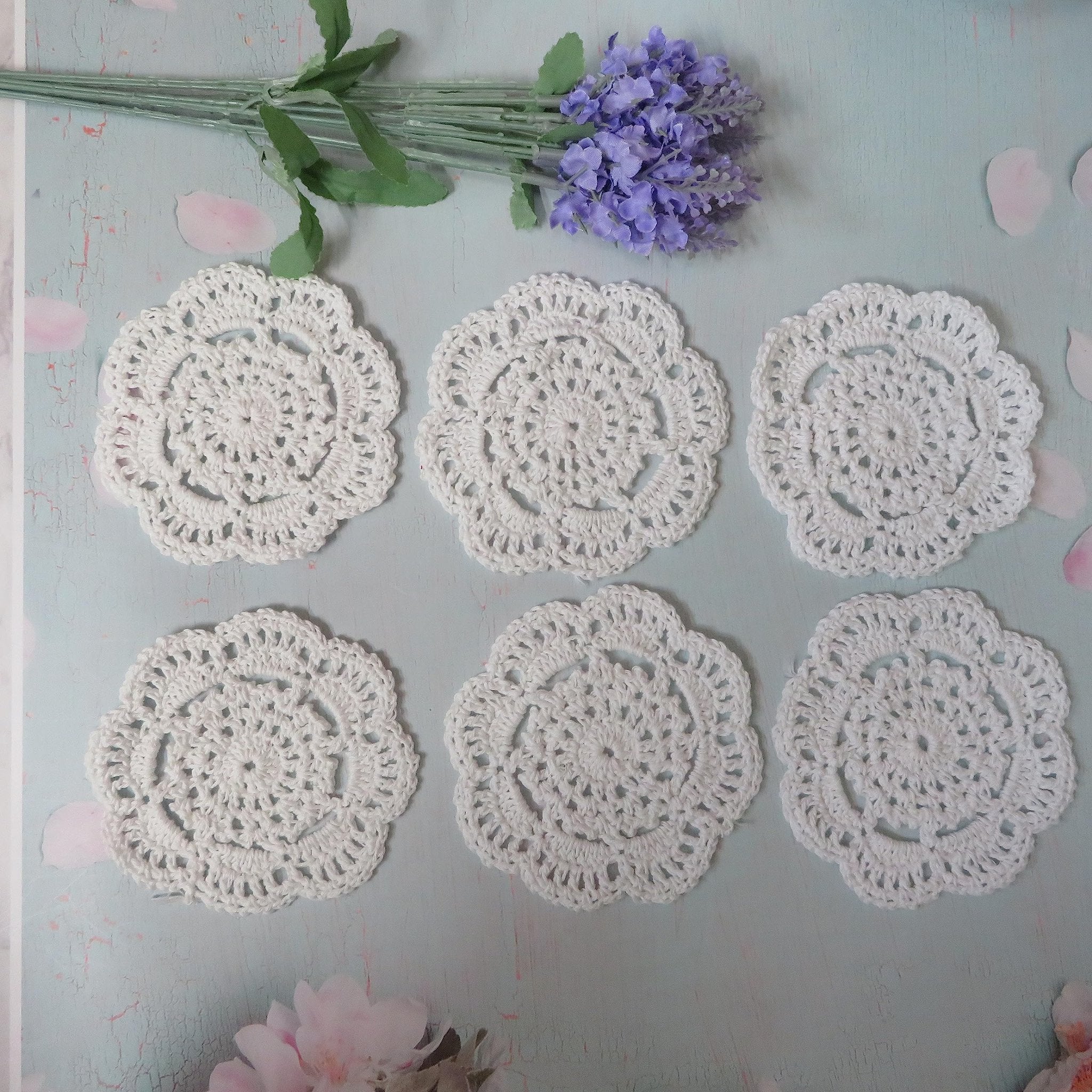 Six white handmade cotton crochet doilies arranged on a light blue distressed background with lavender and pink floral accents.