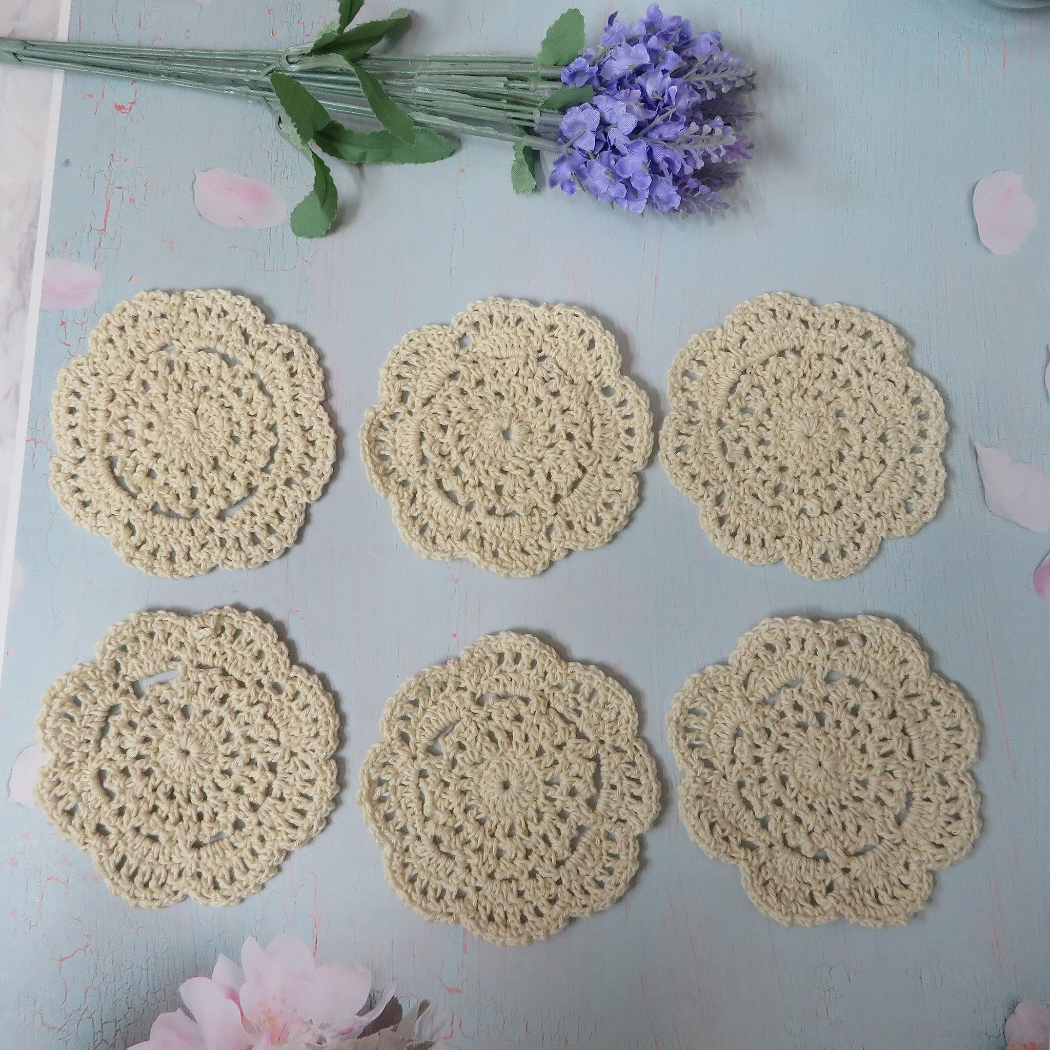 Six cream-colored handmade crochet doilies arranged on a light blue distressed surface with lavender and pink floral accents.