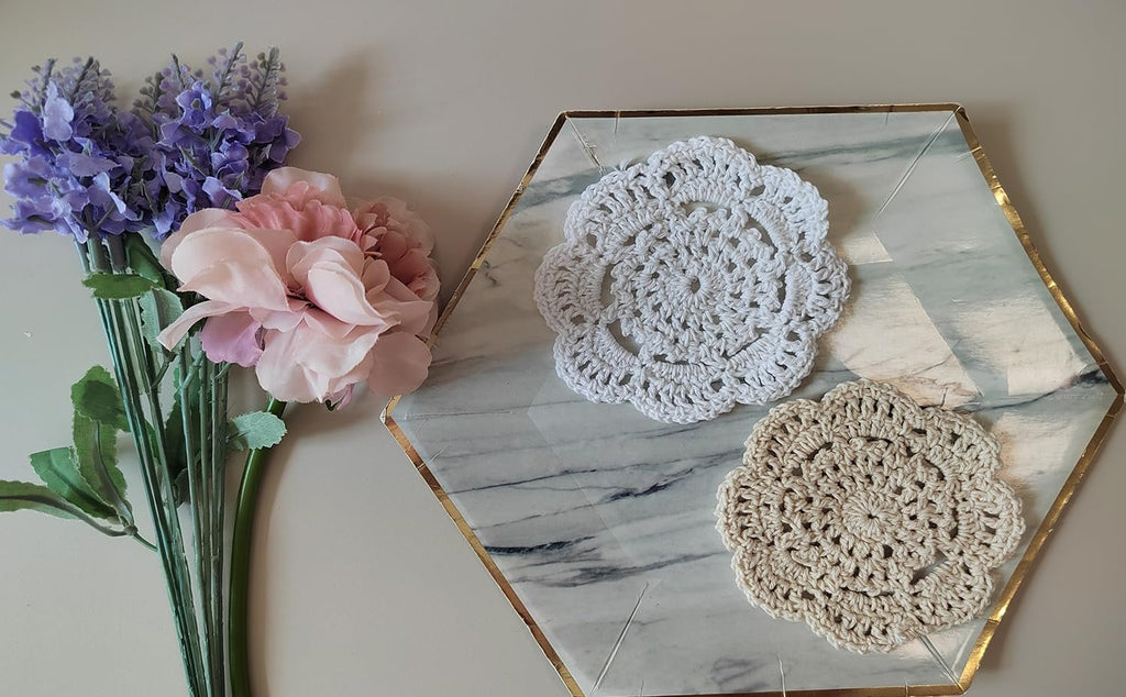 Two handmade crochet doilies, one white and one beige, on a marble-patterned hexagonal tray with artificial flowers.