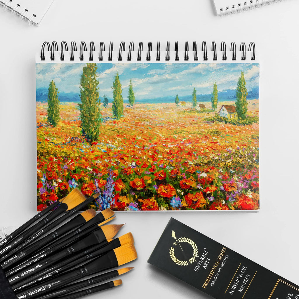 Set of 12 paint brushes with black handles and yellow bristles, arranged next to a spiral-bound sketchbook featuring a landscape painting.