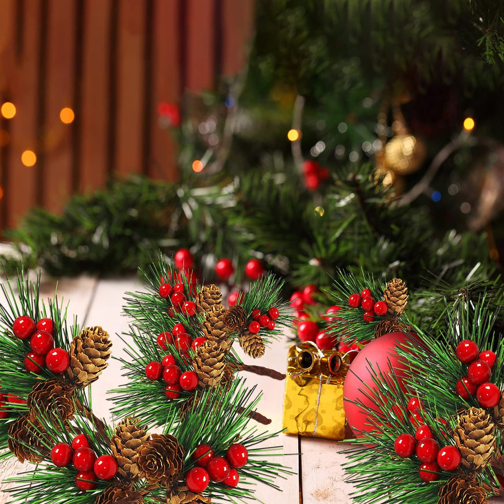 Artificial Christmas pine branches with red berries, pine cones, a gold gift box, and a red ornament on a white wooden surface with a blurred Christmas tree background.
