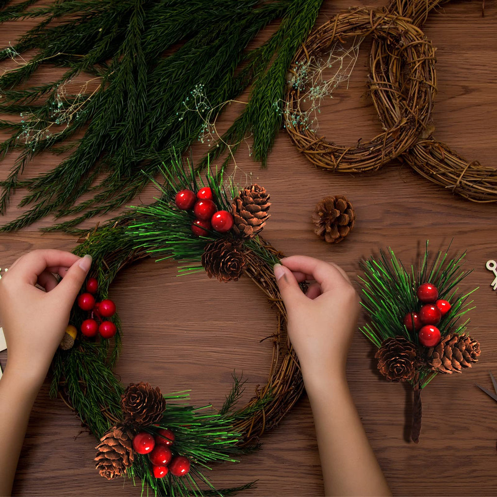 Hands assembling a Christmas wreath with pine branches, red berries, and pine cones on a wooden table.