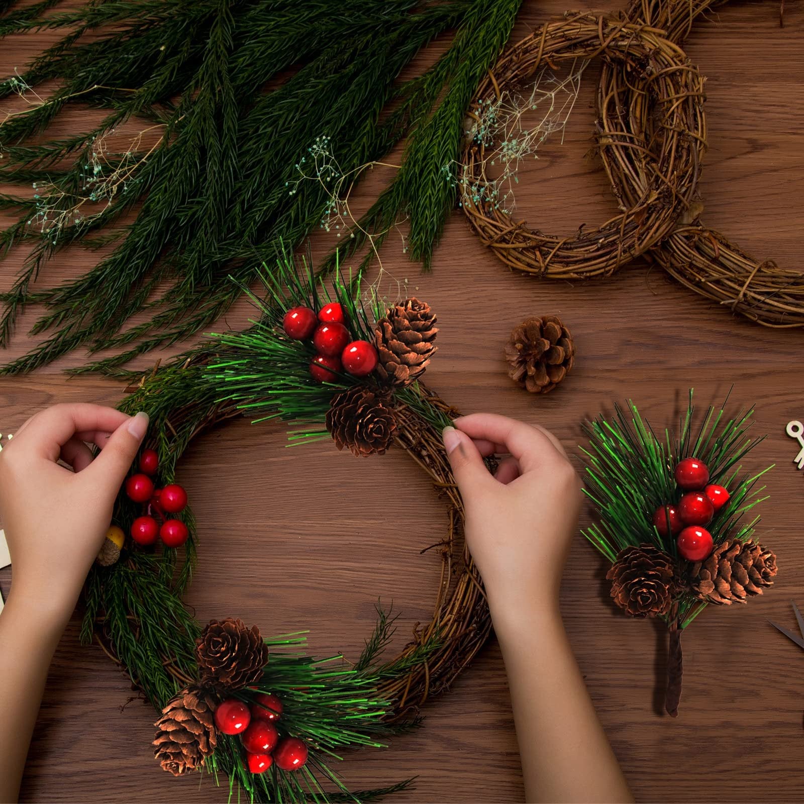 Hands assembling a Christmas wreath with pine branches, red berries, and pine cones on a wooden table.