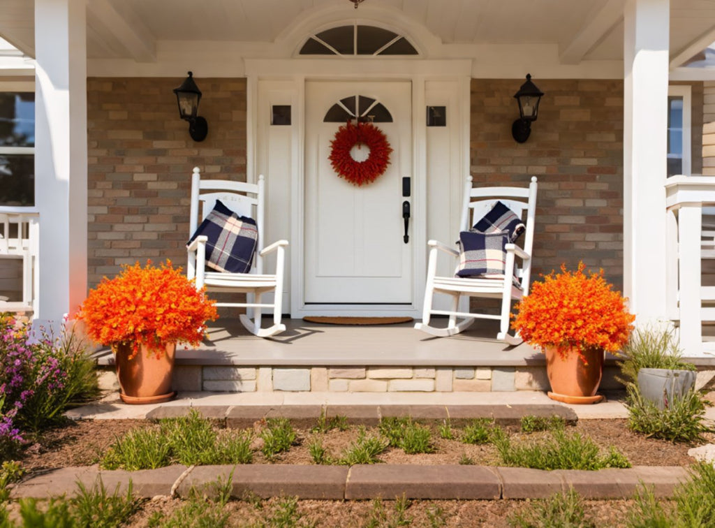 Front porch decorated for fall with two white rocking chairs, orange potted plants, and a red wreath on the door.